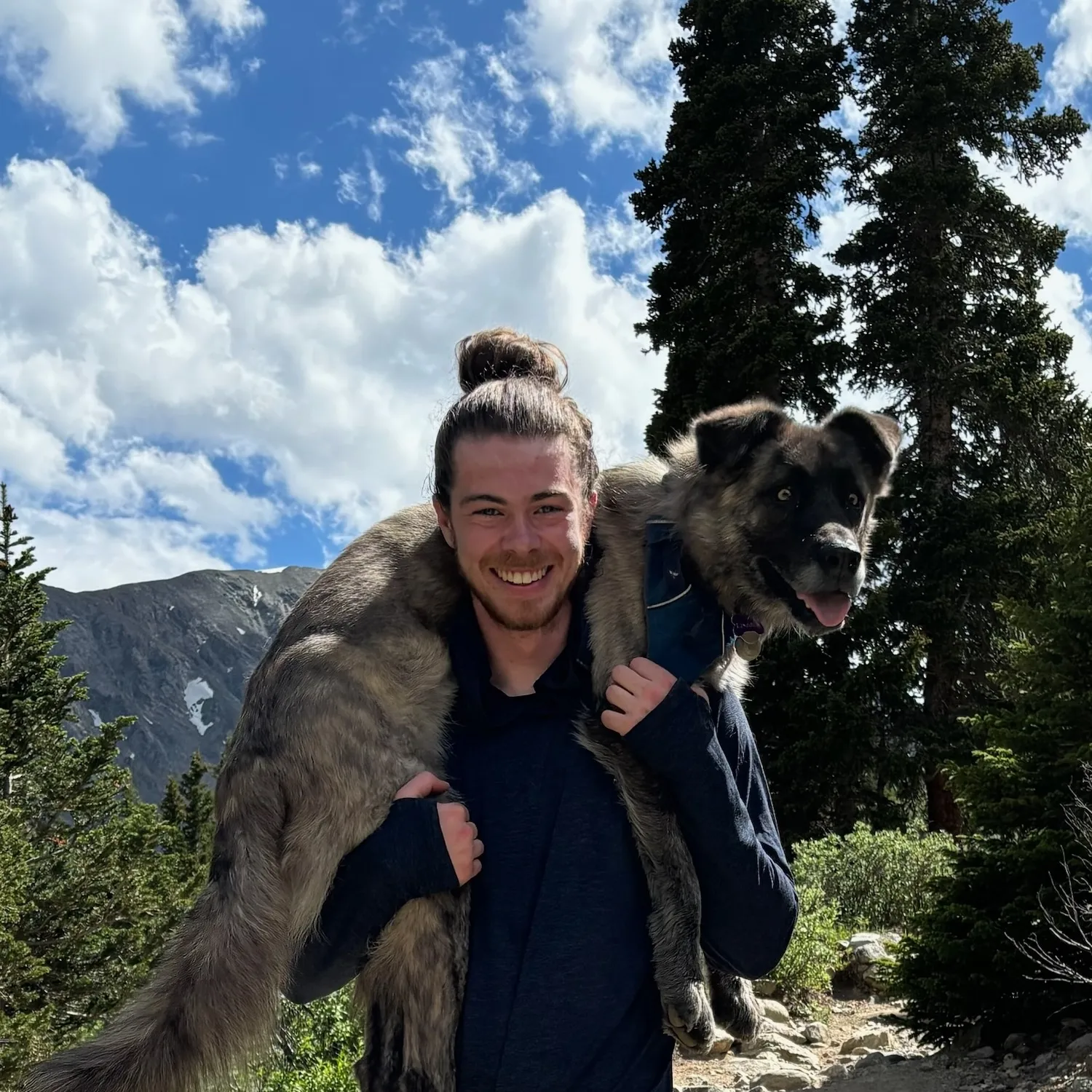 Ethan standing with his late dog, Lincoln, over his shoulders (in a fireman carry). The background is a scenic summer day with a dirt trail, mountain peaks, trees, and other rocks and shrubs too.