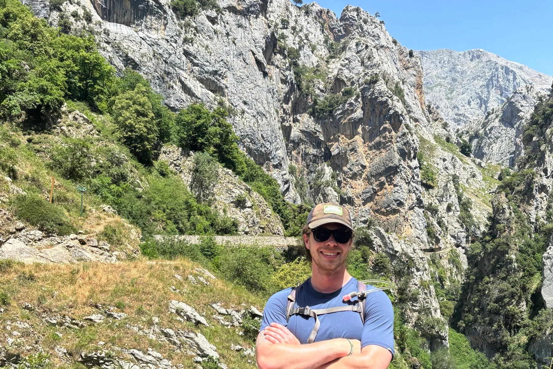Ethan standing with his arms crossed in front of a dramatic mountain gorge with steep cliffs, green slopes, a distant peak, and blue sky on a sunny day. He is wearing sunglasses, a cap, and a hydration vest.